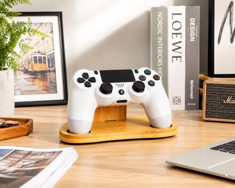 May include: A white wireless game controller rests on a light-colored wooden stand. The controller features black analog sticks, a central touchpad, and standard action buttons including a triangle, circle, cross, and square in their respective colors.