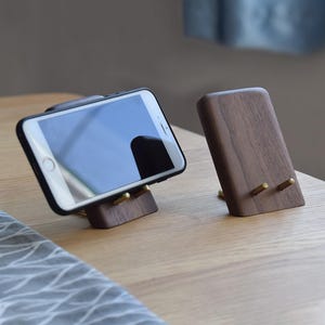 May include: Two wooden phone stands with brass pegs. One stand holds a smartphone with a black case. The stands are made of dark wood and are angled for optimal viewing. The stands are on a light-colored wooden surface.