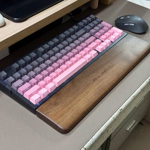 Könnte beinhalten: Eine mechanische Tastatur mit einem Farbverlauf-Tastenkappendesign, das von Dunkelgrau zu Rosa übergeht. Es enthält eine hölzerne Handgelenkauflage mit dem Text "All-Is-Well". Eine kabellose Maus befindet sich rechts.