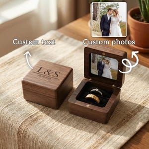 May include: A wooden ring box with a hinged lid, engraved with "L&S" and the date "05.20.2025". The box is open, revealing two rings on a black velvet interior. A small photo is visible inside the lid.