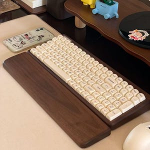 May include: A wooden keyboard with off-white keycaps. The keyboard is set in a dark brown wooden frame with a matching wrist rest. The keys are labeled with black lettering. A phone with a cartoon design is visible in the background.