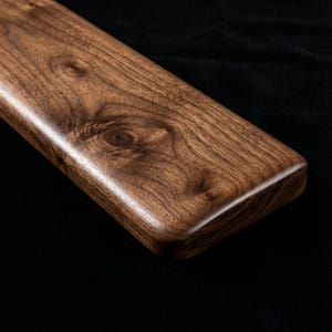 Può includere: Primo piano di un tagliere rettangolare in legno con bordi arrotondati. Il tagliere è realizzato in legno marrone scuro con un motivo a venature visibile e una finitura liscia e lucida. Il tagliere è posto su uno sfondo nero.