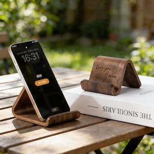 May include: Wooden phone stand with a curved design, holding a smartphone displaying the time. A second stand with "Love you mom" engraved on it sits on a book. The stands are brown and sit on a wooden table.