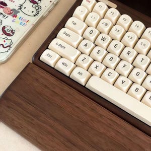 May include: Close-up of a cream-colored mechanical keyboard with a dark wood frame. The keycaps are off-white with black lettering. A phone case with a Hello Kitty design is visible in the background.