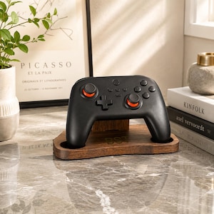 May include: A black wireless video game controller with orange accents on the analog sticks, resting on a dark brown wooden stand. The stand features a smooth, curved base with a small engraved monogram visible on the front center surface.