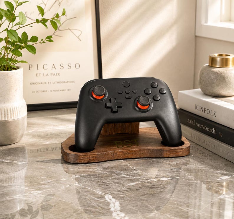 May include: A black wireless video game controller with orange accents on the analog sticks, resting on a dark brown wooden stand. The stand features a smooth, curved base with a small engraved monogram visible on the front center surface.