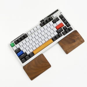 Peut inclure: Un clavier mécanique blanc avec des touches noires, blanches, rouges, bleues et jaunes. Le clavier est accompagné d'un repose-poignet en bois de chaque côté. Le clavier porte le texte "Kzzi" en haut.