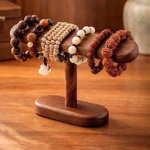 May include: A wooden bracelet display stand holding several beaded bracelets. The bracelets are various colors and textures, including brown, beige, and white. The stand is made of dark wood and has a simple, elegant design.