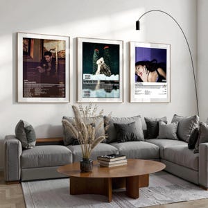 May include: Three framed posters with album art hang above a gray sectional sofa. The posters feature images of musicians and album details. A round wooden coffee table and decorative vase with dried plants complete the living room decor.