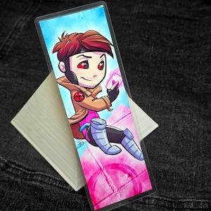 Pode incluir: Um marcador de página com uma ilustração de desenho animado do Gambit dos X-Men. Ele está vestindo uma jaqueta marrom, uma camisa rosa e calças azuis. Ele está segurando um cartão de jogo rosa na mão. O fundo é rosa e branco.