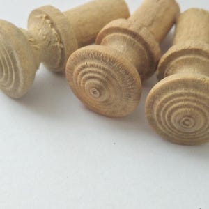 May include: Four unfinished wooden knobs with a light brown color. Each knob has a rounded, grooved base and a cylindrical top. The knobs are arranged on a white surface.