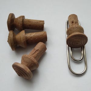 Peut inclure: Quatre boutons en bois avec un dessus arrondi et une base cylindrique. Les boutons sont de couleur marron et ont une finition en bois naturel. Les boutons sont disposés en demi-cercle sur une surface blanche. Un trombone argenté maintient l'un des boutons.