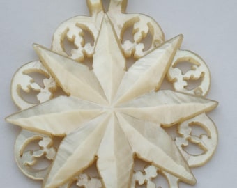May include: A cream-colored pendant with a starburst design. The pendant features a central eight-pointed star and an outer ring with decorative cut-out patterns. A small, circular metal loop is attached at the top for hanging.