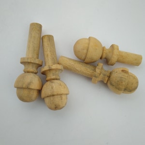 May include: Several unfinished wooden finials are displayed, each with a rounded base, a decorative central section, and a cylindrical top. The natural wood grain is visible. They are arranged on a white surface.