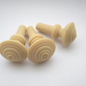 May include: Three beige plastic push pins with a circular, ridged top. The push pins are designed for use with cork boards or notice boards.