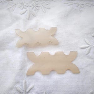 Peut inclure: Deux outils de coiffure en plastique translucide pâle. Chaque outil a une forme incurvée avec trois découpes arrondies et deux extensions incurvées. Les outils sont sur un tissu blanc texturé avec des détails floraux brodés.