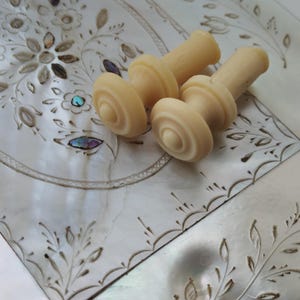 May include: Three ivory-colored cabinet knobs with a decorative, circular design. The knobs are on a white surface with a floral pattern.