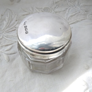 May include: A small, clear glass jar with a faceted design and a shiny, silver lid. The lid has markings on it. The jar is sitting on a white, patterned surface. The jar is likely for storing small items.