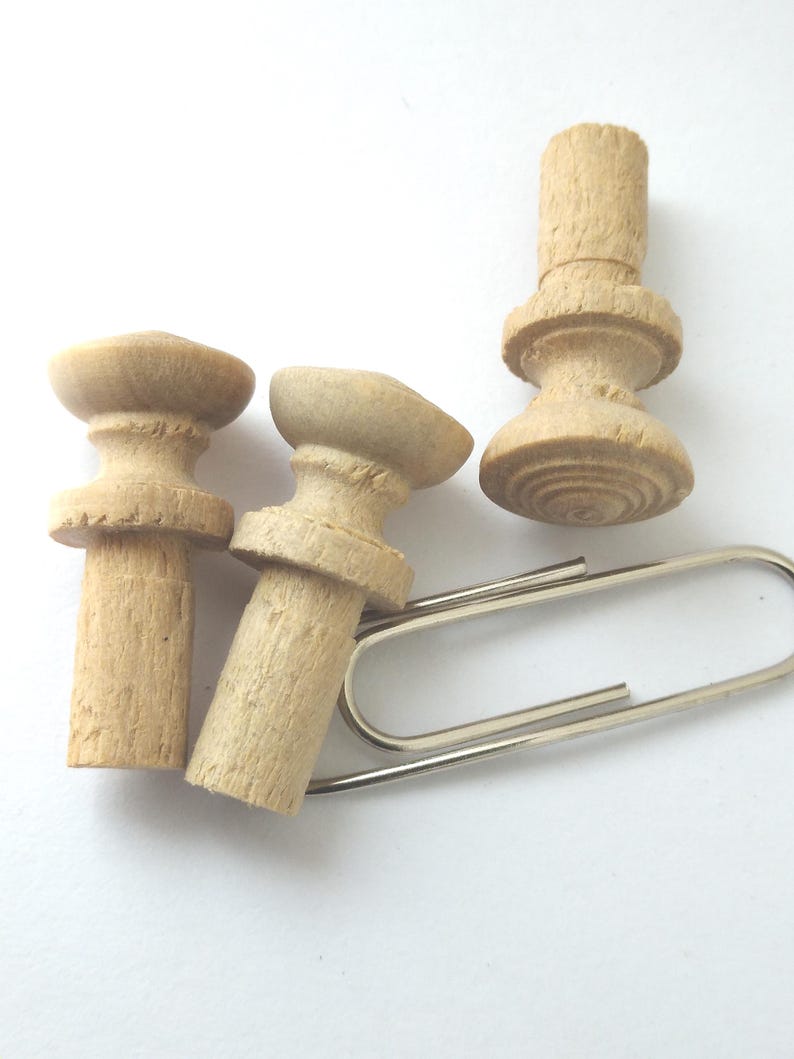 May include: Three unfinished wooden bottle stoppers with a decorative design, next to a silver paperclip. The stoppers are light brown and have a textured surface. The paperclip is made of shiny metal.
