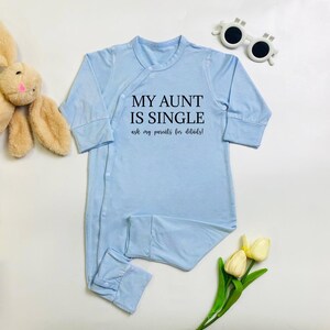 May include: A light blue baby onesie with the text "MY AUNT IS SINGLE ask my parents for details!". The onesie has long sleeves and snaps down the front. A plush bunny and sunglasses are also in the image.