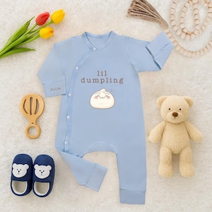 May include: A light blue baby onesie with the text "lil dumpling" and a dumpling graphic. The onesie is surrounded by a wooden rattle, a teddy bear, and blue bear slippers. A bouquet of tulips is also present.