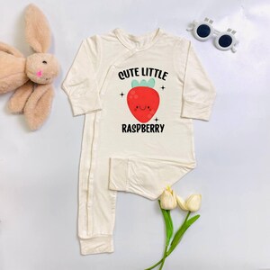 May include: Cream-colored baby onesie with long sleeves and attached feet. The front features a red raspberry graphic with the text "Cute Little Raspberry". The onesie has a wrap-style closure and is made of a soft material.