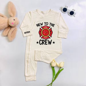 May include: Cream-colored baby sleeper with long sleeves. The front features a red and yellow firefighter emblem with the text "NEW TO THE FIRE DEPT. CREW". The name "HAYDEN" is printed on the sleeve. A plush bunny and sunglasses are in the background.