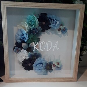 May include: A framed floral arrangement featuring a variety of blue, teal, and white artificial flowers. The name "KODA" is displayed in white script across the center of the clear glass. The frame is made of light-colored wood.