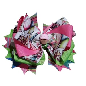 May include: A large, layered hair bow featuring a cupcake embellishment. The bow incorporates bright pink, green, and striped ribbons. Some ribbons have a white background with birthday-themed illustrations, including balloons and party hats.