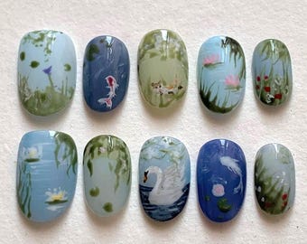 May include: A set of ten painted artificial nails. Each nail features a different miniature landscape design, including koi fish, swans, and water lilies, in shades of blue, green, and white. The nails are oval-shaped and arranged on a white surface.