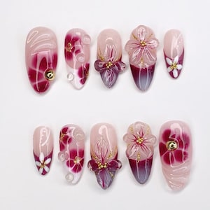 Op de afbeelding: Een set van tien kunstnagels met diverse ontwerpen. De nagels hebben een combinatie van roze, wit en bordeauxrode kleuren, met bloemen- en abstracte patronen. Sommige nagels hebben 3D bloemenversieringen en gouden accenten.