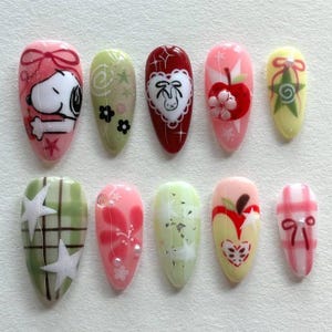 Könnte beinhalten: Ein Satz von zehn künstlichen Nägeln mit verschiedenen Designs. Die Nägel zeigen Cartoonfiguren, Äpfel, Herzen und florale Muster in Rosa, Grün, Rot und Gelb. Einige Nägel haben Glitzer und Sternakzente.