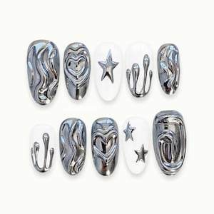 Peut inclure: Un ensemble de dix faux ongles de différentes formes. Certains ongles sont argentés avec un aspect de métal liquide. D'autres sont blancs avec des motifs de cœur, d'étoile et de goutte argentés. Les ongles sont de forme ovale.