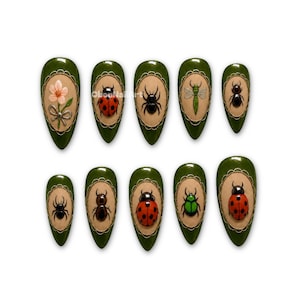 Op de afbeelding: Een set van tien amandelvormige press-on nagels met een donkergroene basis en een centraal beige ovaal kader. Elke nagel heeft een verhoogd 3D-ontwerp met een roze bloem, lieveheersbeestjes, spinnen en diverse kevers in rood, groen en zwart.