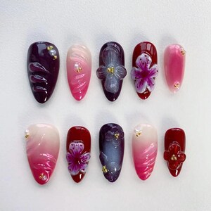 Peut inclure: Dix faux ongles en forme d'amande. Les ongles présentent divers motifs, notamment des effets ombrés, des décorations florales et des accents 3D. Les couleurs comprennent des nuances de rose, de rouge et de violet, avec des accents dorés.