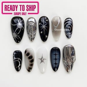 Op de afbeelding: Een set van tien kunstnagels met diverse ontwerpen in zwart, wit en metallic zilver. De patronen omvatten sterren, wervelingen, gradiënten en rastermotieven, waarbij sommige nagels verhoogde 3D-metaaltexturen en kleine zilveren studs bevatten.