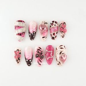 Peut inclure: Un ensemble de dix faux ongles avec divers motifs. Les ongles présentent des couleurs rose, blanc et noir, avec des embellissements tels que des strass, des nœuds et des breloques en forme de cœur. Certains ongles ont un imprimé léopard, d'autres des motifs abstraits.