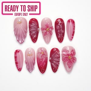 Può includere: Un set di dieci unghie finte a mandorla in tonalità rosa e rosso scuro. I design presentano rilievi floreali 3D, texture in rilievo simili a conchiglie, piccole perline dorate e perle bianche. Alcune unghie hanno una finitura sfumata liscia, mentre altre mostrano dettagli scolpiti complessi e superfici lucide. Il testo QbonNailArt è visibile su un'unghia.