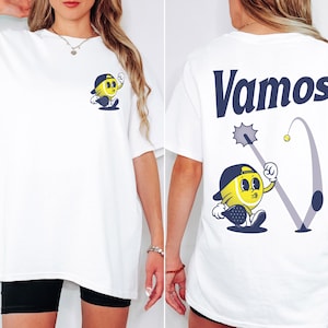 May include: White t-shirt with a cartoon illustration of a yellow tennis ball character wearing a hat and holding a racket on the front. The back of the shirt has the word "Vamos" in blue text and a cartoon illustration of a tennis ball character hitting a ball with a racket.
