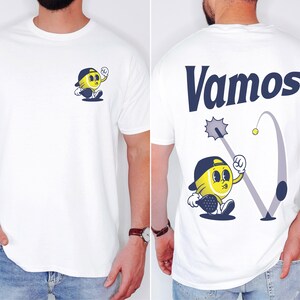 May include: White t-shirt with a cartoon tennis ball character wearing a hat and holding a racket on the front. The back of the shirt has the word "Vamos" in blue lettering and a cartoon tennis ball character swinging a racket.