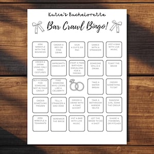 Pode incluir: Um cartão de jogo em papel branco intitulado Katie's Bachelorette Bar Crawl Bingo com texto preto e ilustrações de laços. A grelha contém vinte e cinco quadrados com várias atividades e um ícone de anel de diamante no centro.