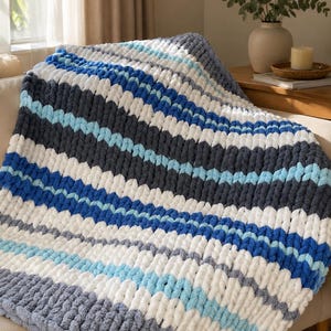 Könnte beinhalten: Eine grob gestrickte Decke mit einem wiederkehrenden Streifenmuster in den Farben Dunkelgrau, Weiß, Hellblau und leuchtendem Blau. Die Decke besteht aus dicken, weichen Garnschlaufen, die eine strukturierte und flauschige Oberfläche erzeugen.