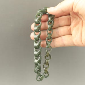 May include: A dark green chain-link bracelet with a clasp, held against a gray background. The bracelet's links are a muted, olive-toned green, and the clasp is a slightly lighter shade. The design features a series of interlocking, rounded links.