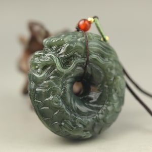Könnte beinhalten: Dunkelgrüner Jade-Anhänger in Form eines gewundenen Drachen. Der Drache hat detaillierte Schnitzereien an Kopf und Körper. Eine rote Perle und eine grüne Kordel sind befestigt. Der Anhänger hat einen Durchmesser von etwa 5 cm.