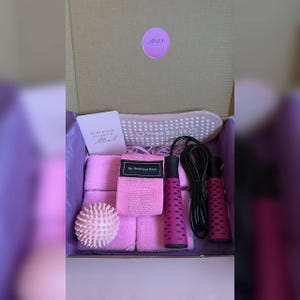 Puede incluir: Una caja de regalo de fitness rosa que contiene una bola de masaje, una banda de resistencia, una cuerda para saltar, calcetines de yoga y dos toallas pequeñas. La caja también incluye una tarjeta con el texto "Be the energy you want to Attract".