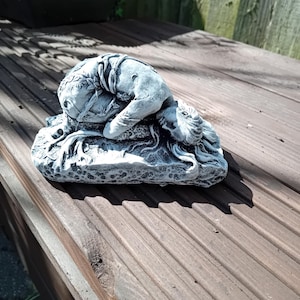 May include: A grey concrete sculpture of a woman lying on her side with her head resting on her hand. The sculpture is on a rectangular base with a textured surface.