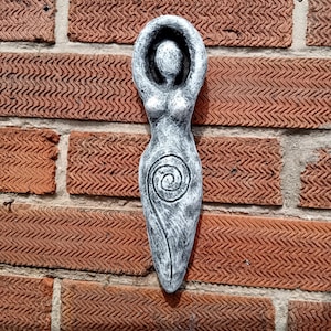 May include: A white ceramic sculpture of a female figure with raised arms and a spiral design on the torso. The sculpture is mounted on a brick wall.