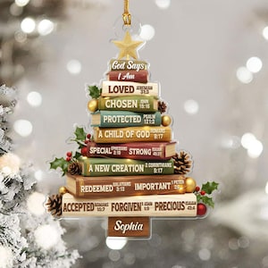 May include: A Christmas tree ornament shaped like a stack of books, with a gold star on top. The books have phrases like "Loved," "Chosen," and "Precious." The name "Sophia" is at the bottom. Holly, berries, and pinecones are included.