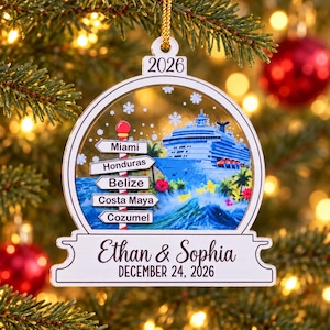 K&ouml;nnte beinhalten: Ein wei&szlig;er, kugelf&ouml;rmiger Weihnachtsbaumschmuck mit einem Kreuzfahrtschiff, tropischen Pflanzen und einem Wegweiser mit der Aufschrift Miami, Honduras, Belize, Costa Maya und Cozumel. Der Schmuck zeigt die Namen Ethan & Sophia und 24. Dezember 2026.