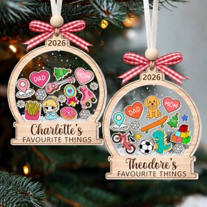 May include: Two wooden snow globe ornaments with red and white gingham bows and white ribbons. One features the text Charlotte's FAVOURITE THINGS 2026 with icons of hearts, a guitar, fries, and a roller skate. The other says Theodore's FAVOURITE THINGS 2026 with a dog, skateboard, soccer ball, and dinosaur.